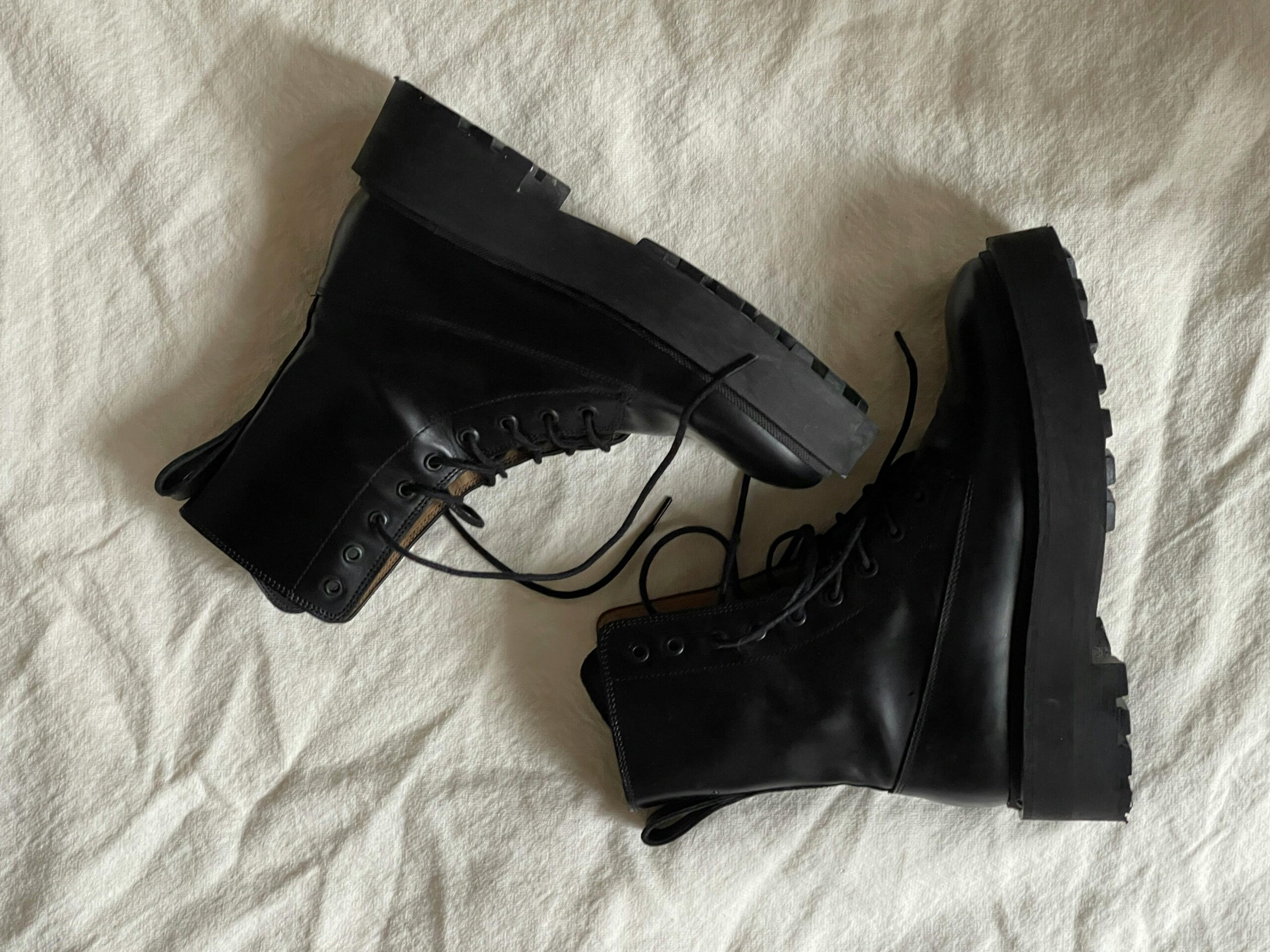 Pair of black lace-up leather work boots with thick treaded soles, lying on a crumpled white fabric background.